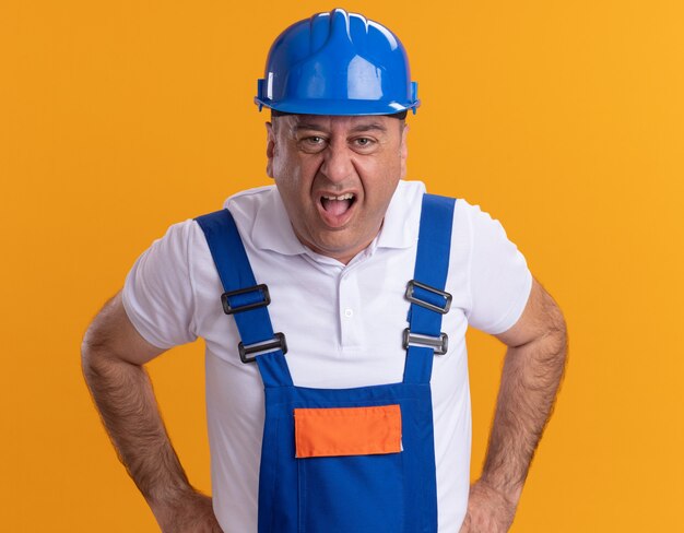 オレンジ色の壁に隔離された正面を見て制服を着たイライラした大人のビルダー男