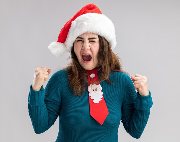 Annoyed adult caucasian woman with santa hat and santa tie keeping fists isolated on white background with copy space