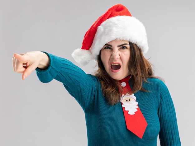 Annoyed adult caucasian woman with santa hat and santa tie pointing at side isolated on white wall with copy space