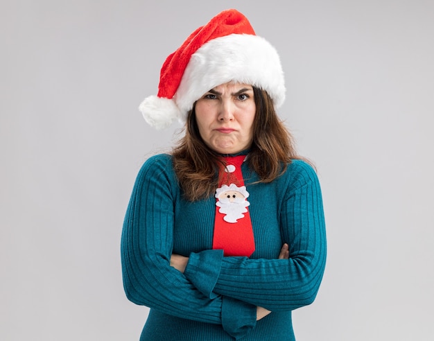 Annoyed adult caucasian woman with santa hat and santa tie standing with crossed arms isolated on white wall with copy space