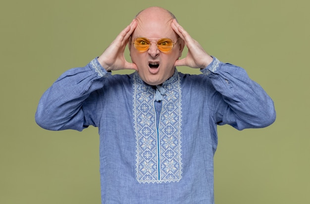 Free photo annoyed adult slavic man in blue shirt wearing sunglasses holding his head