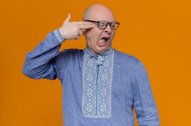 Annoyed adult slavic man in blue shirt and with optical glasses keeping hand on his temple gesturing gun sign