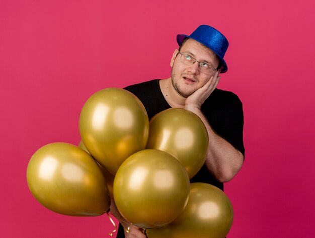 Annoyed adult slavic man in optical glasses wearing blue party hat puts hand on ear and holds helium balloons looking at side 