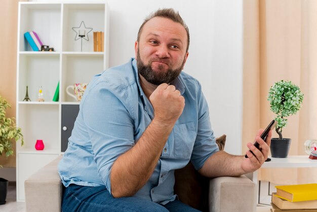 Annoyed adult slavic man sits on armchair keeping fist and holding phone inside the living room