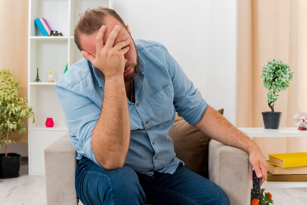 Annoyed adult slavic man sits on armchair putting hand on face holding tv remote inside the living room