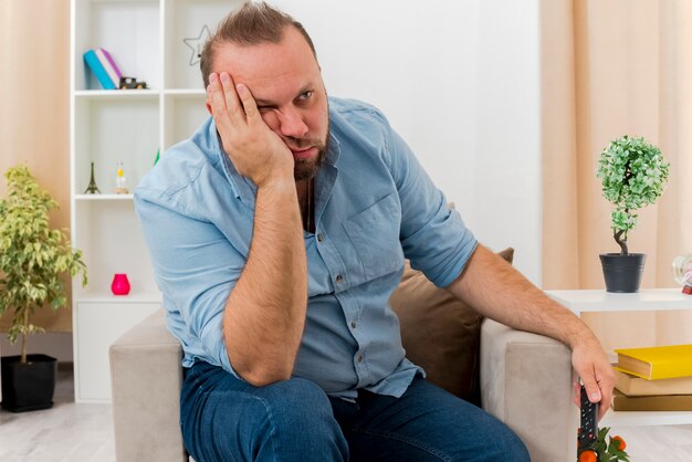 Annoyed adult slavic man sits on armchair putting hand on face looking at side inside the living room