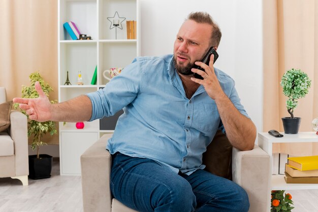 Annoyed adult slavic man sits on armchair talking on phone looking and pointing at side inside the living room