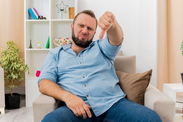 Annoyed adult slavic man sits on armchair and thumbs down inside the living room