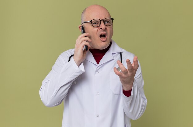 Annoyed adult slavic man with optical glasses in doctor uniform with stethoscope talking on phone looking at side