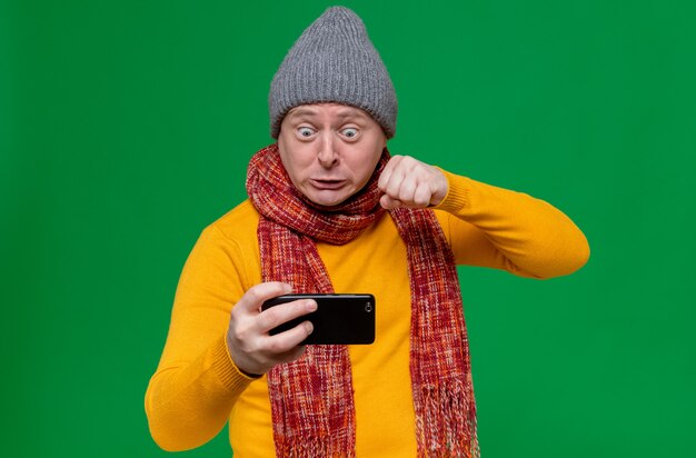 Annoyed adult slavic man with winter hat and scarf around his neck holding phone and keeping fist 