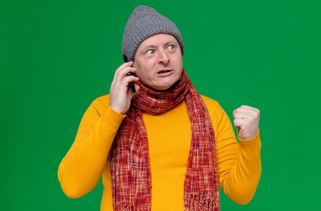 Annoyed adult slavic man with winter hat and scarf around his neck talking on phone and keeping fist looking at side 