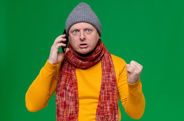 Annoyed adult slavic man with winter hat and scarf around his neck talking on phone and keeping fist