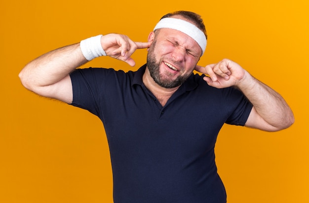 annoyed adult slavic sporty man wearing headband and wristbands closing his ears with fingers isolated on orange wall with copy space