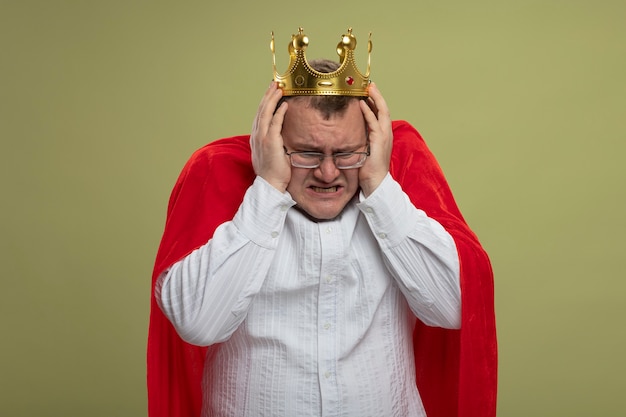 Бесплатное фото Раздраженный взрослый славянский супергерой в красном плаще в очках и короне, держащий голову, смотрит вниз, изолирован на оливково-зеленой стене с копией пространства