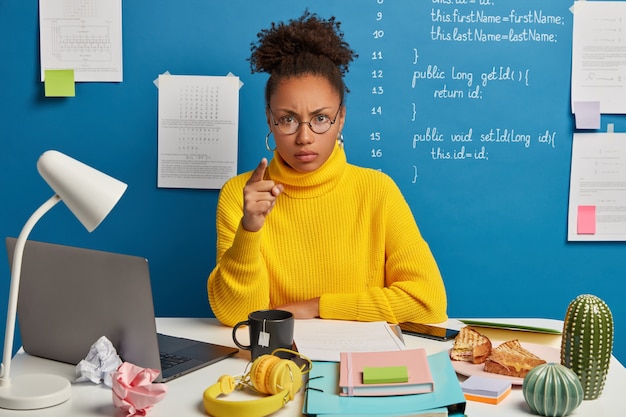 Annoyed Afro American woman worker points at you and blames in doing something wrong, wears round glasses and yellow jumper, sits in coworking space with mess on table.