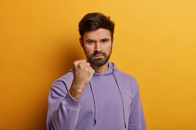 Annoyed aggressive Caucasian man shows fist, looses temper, looks at person with hate, promises to revenge, dressed in violet hoodie, isolated on yellow wall. Negative emotions concept