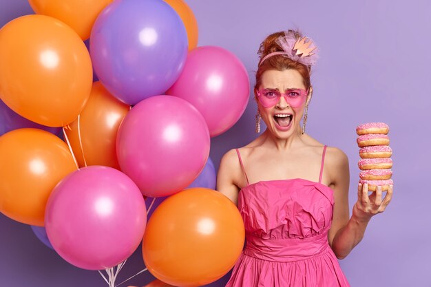 Раздраженная злая рыжая женщина в розовом праздничном платье и солнцезащитных очках в форме сердца держит кучу пончиков, громко восклицает, позирует с разноцветными гелиевыми шарами