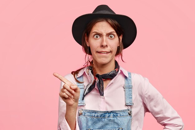 Annoyed angry woman gardener complains at noisy neighbours, points aside in direction where they live, dressed in workwear