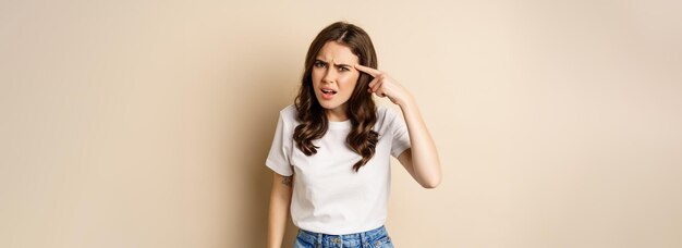 Annoyed and angry woman pointing finger at head scolding someone standing over beige background