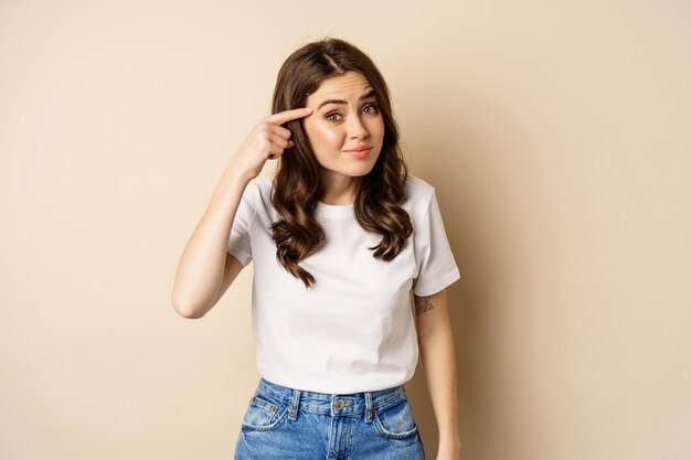 Annoyed and angry woman pointing finger at head, scolding someone, standing over beige background