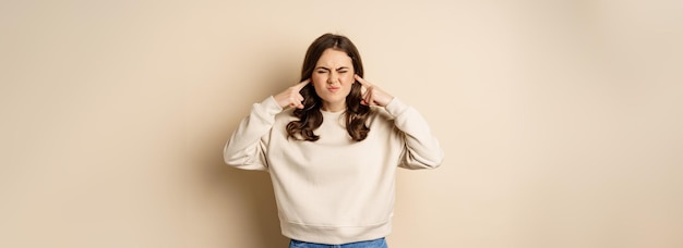 Free photo annoyed and angry woman shut ears complaining at loud noisy music noise standing over beige background