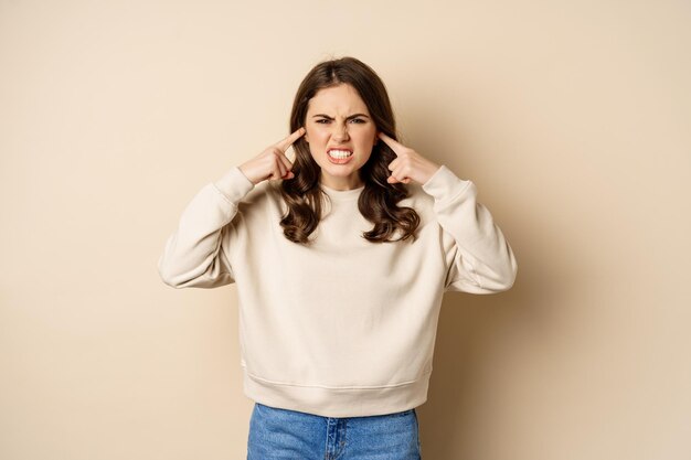 Annoyed and angry woman shut ears complaining at loud noisy music noise standing over beige background
