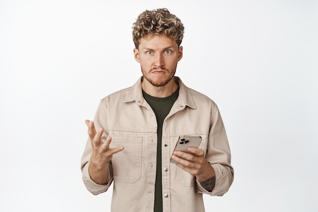 Annoyed and angry young man holding mobile phone looking pissedoff at camera standing frustrated against white background