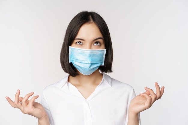 Раздраженная азиатка в медицинской маске пожимает плечами и смотрит вверх с обеспокоенным выражением лица, стоящим на белом фоне
