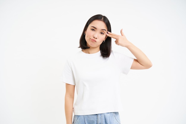 Free photo annoyed asian woman student shows finger pistol near head looks bothered and tired stands isolated o