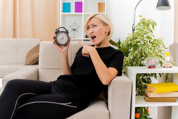 Annoyed beautiful blonde russian woman lying on armchair holding and pointing at alarm clock inside the living room