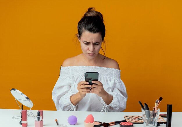 Annoyed beautiful girl sits at table with makeup tools holding and looking at phone isolated on orange wall