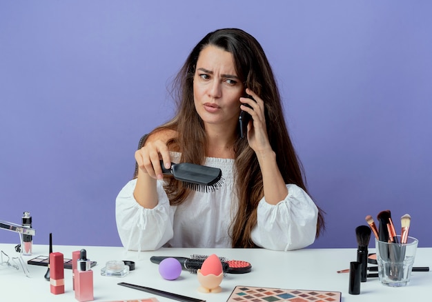 Annoyed beautiful girl sits at table with makeup tools holds hair comb talking on phone looking at side isolated on purple wall