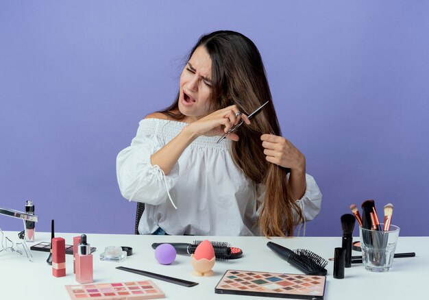 Annoyed beautiful girl sits at table with makeup tools pretends to cut hair with scissors looking at side isolated on purple wall