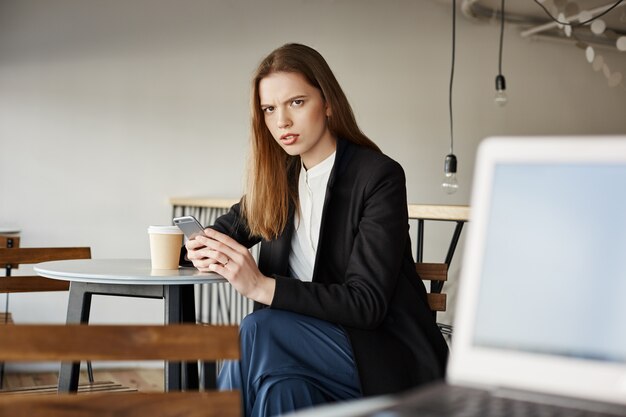 Annoyed businesswoman sit in cafe with mobile phone and looking at person angry