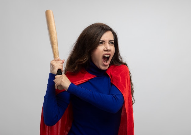 Annoyed caucasian superhero girl with red cape holds baseball bat and looks at side isolated on white wall with copy space