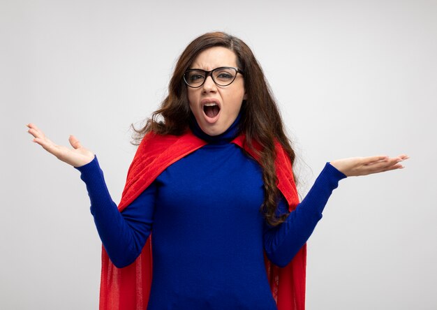 Annoyed caucasian superhero girl with red cape in optical glasses holds hands open on white