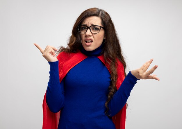 Раздраженная кавказская девушка-супергерой с красной накидкой в оптических очках стоит с поднятой рукой