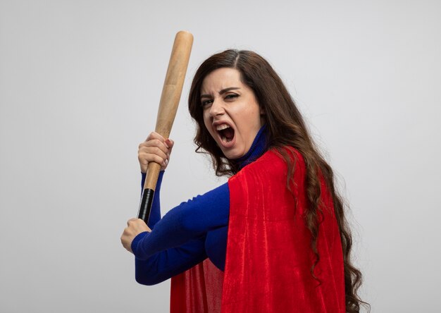 Annoyed caucasian superhero girl with red cape stands sideways and holds baseball bat isolated on white wall with copy space