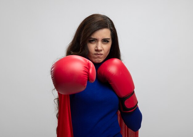 Раздраженная кавказская девушка-супергерой в красной накидке в боксерских перчатках делает вид, что бьет, изолирована на белой стене с копией пространства