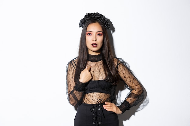 Annoyed and confused asian woman in witch halloween costume pointing at herself, looking bothered and unamused, standing reluctant over white background.