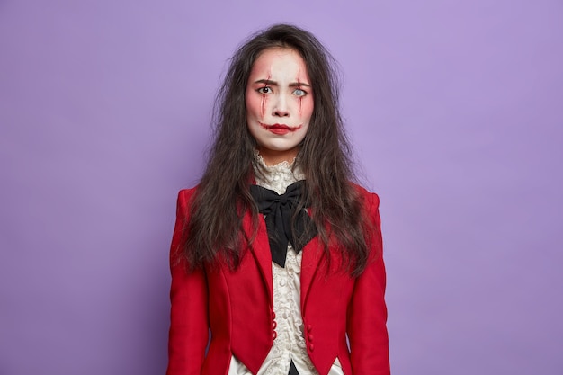 Annoyed displeased brunette woman has image of scary monster celebrates October festival and halloween wears professional spooky makeup poses against purple wall