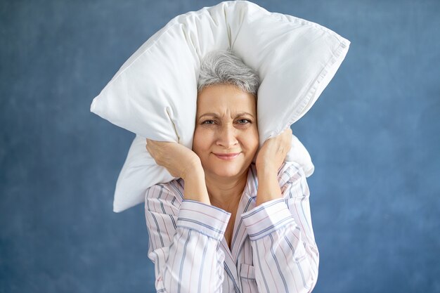 Annoyed dissatisfied mature woman with gray hair grimacing having sleepless night because of loud music