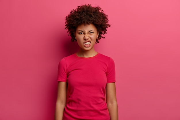 イライラした不満の女性は、歯を食いしばってイライラし、不快に見え、カジュアルなtシャツを着て、ピンクの壁に向かって屋内でポーズをとります。否定的な表情の概念