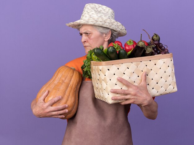 Раздраженная пожилая женщина-садовник в садовой шляпе держит корзину с овощами и тыкву, глядя в сторону, изолированную на фиолетовой стене с копией пространства