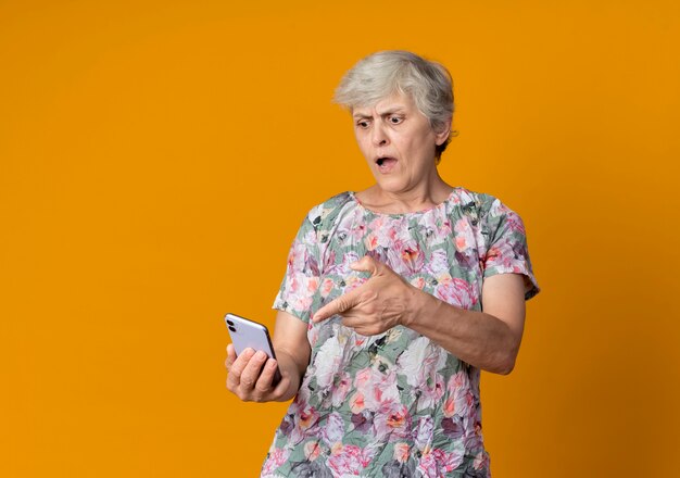 Annoyed elderly woman holds and points at phone isolated on orange wall