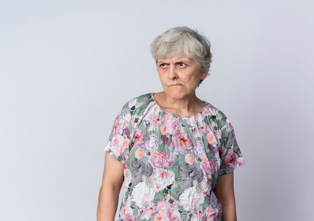 Раздраженная пожилая женщина стоит, глядя в сторону, изолированную на белой стене