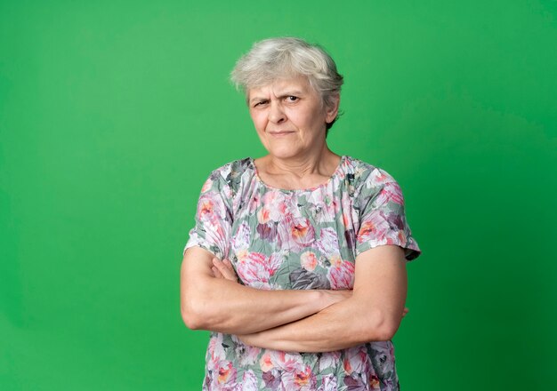 Annoyed elderly woman stands with crossed arms isolated on green wall