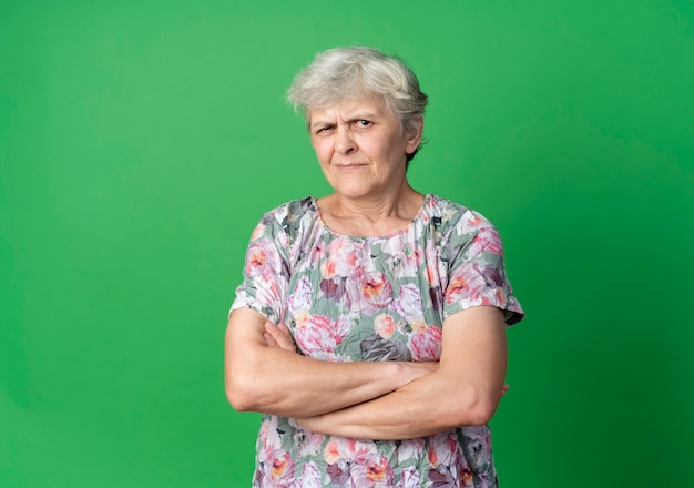 Раздраженная пожилая женщина стоит со скрещенными руками на зеленой стене