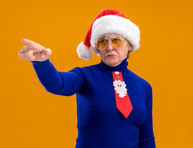 annoyed elderly woman in sun glasses with santa hat and santa tie looking and pointing at side isolated on orange background with copy space