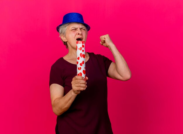 Annoyed elderly woman wearing party hat holds confetti cannon and keeps fist with closed eyes on pink